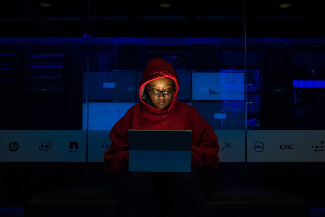 Focused woman in a red hoodie using a laptop in a dimly lit room with technology brands displayed.